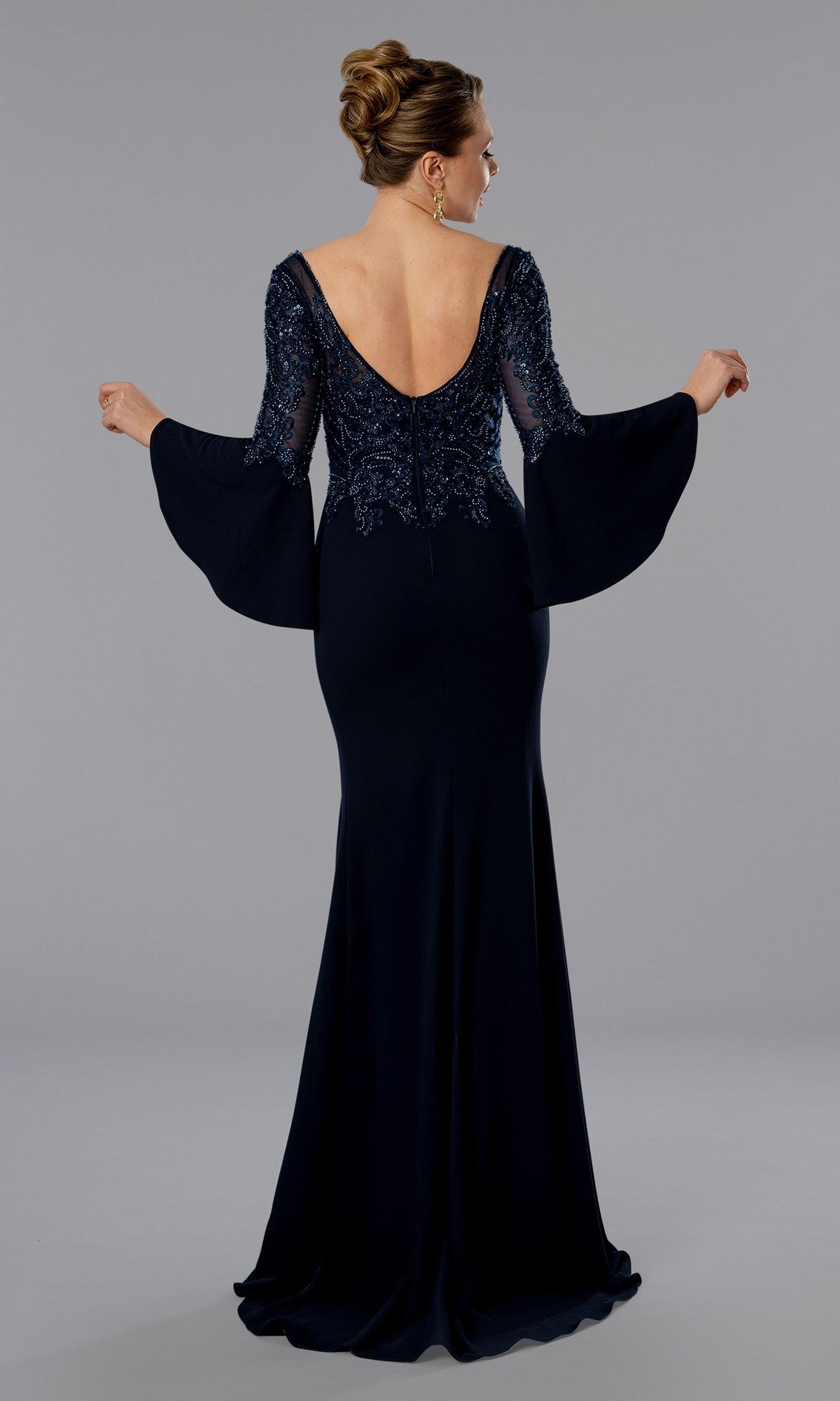A woman stands against a plain grey background wearing the Stella Couture 25366, a long formal evening gown in navy blue with flared sleeves, elegant lines, and an open back as she faces away from the camera.